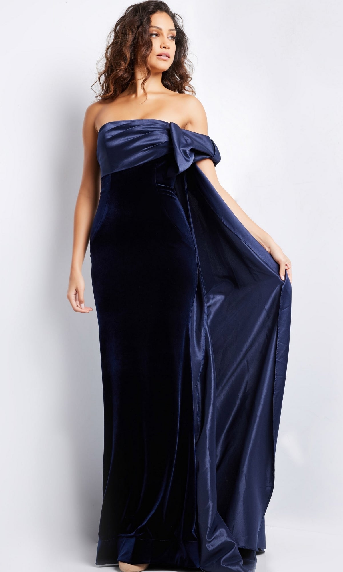 A woman wearing the Long Prom Dress 37391 by Jovani, a navy blue velvet fitted gown featuring a satin draped detail on one side, stands against a plain light background.